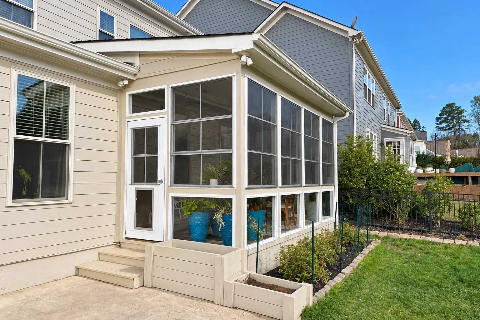A screened patio enclosure with white framing and a single door extends from a light-colored house beside a small garden.