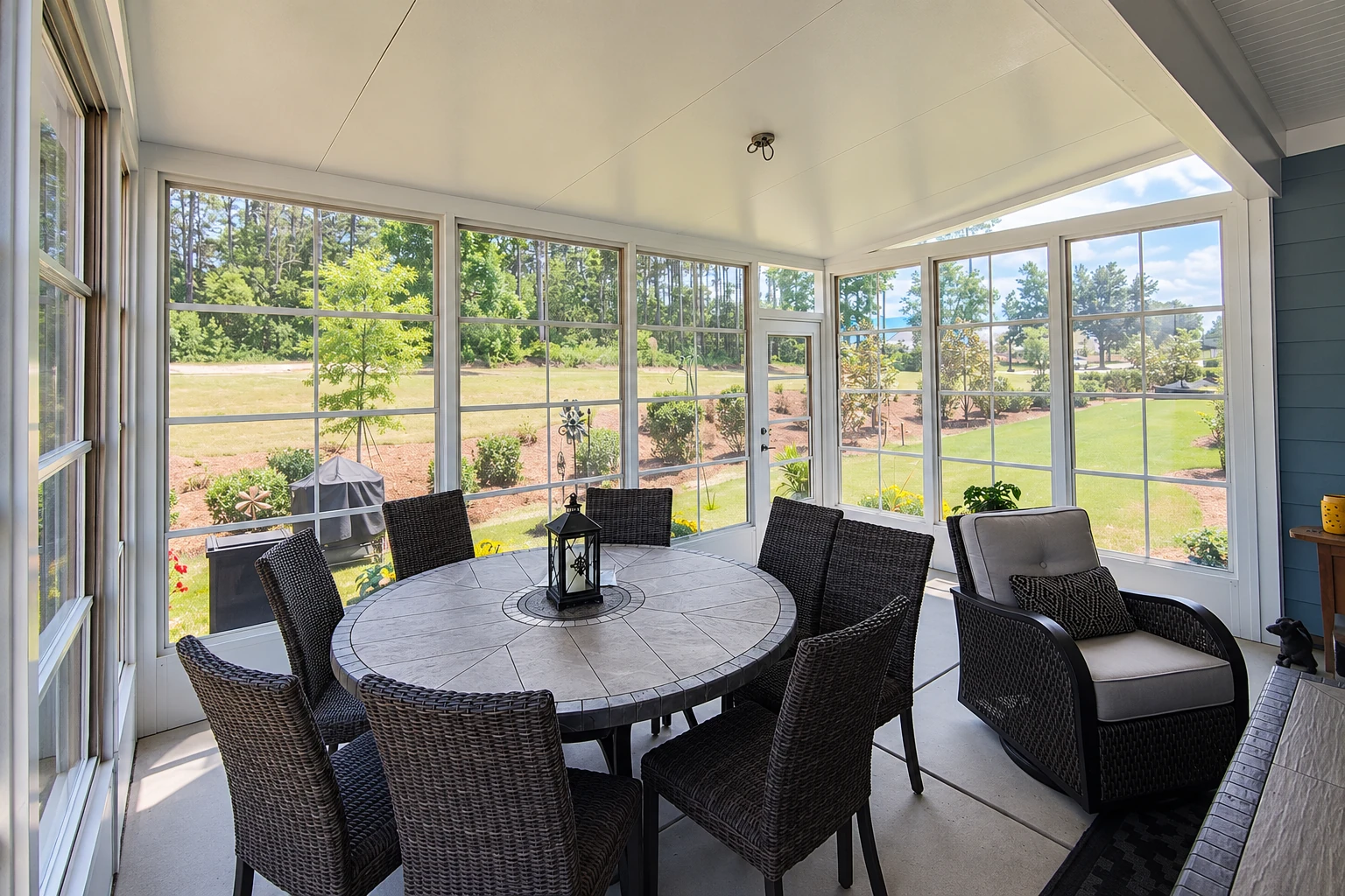 Bright sunroom addition with white-framed windows, a round dining table, and dark wicker chairs overlooking a lawn.